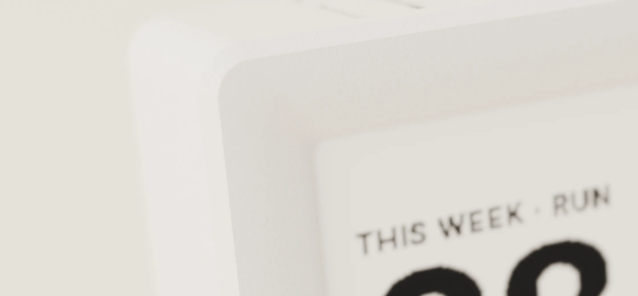 A close view of the display’s top-left corner: the matte sintered-nylon shell and its rounded edge, with the panel’s “THIS WEEK · RUN” line and the top of a large numeral at the edge of the frame.